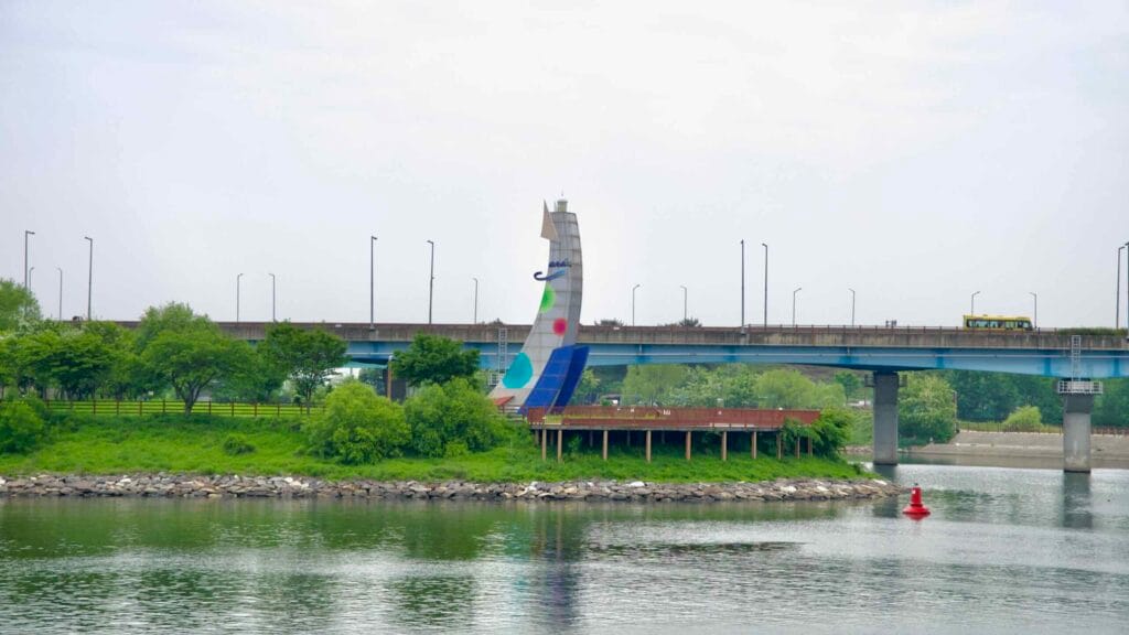 A stylized sail-shaped sculpture marks the riverside deck at Duri Ecological Park, set beside a traffic bridge and framed by greenery along the Ara Waterway.