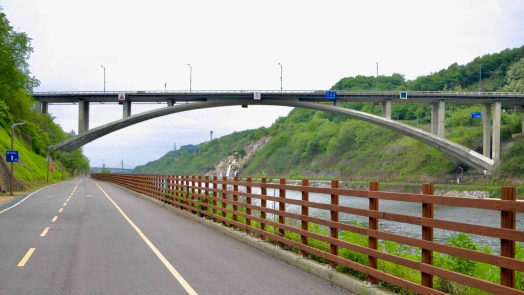 Moksang Bridge spans the Gyeongin Ara Waterway as the Ara Bicycle Path runs beneath it, framed by safety rails, spring foliage, and distant bridges further down the canal.
