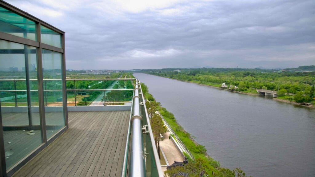 From a rooftop observation platform above Baekseok Bridge, the Ara Canal stretches westward beneath cloudy skies, flanked by parkland, tree-lined roads, and riverside pavilions.