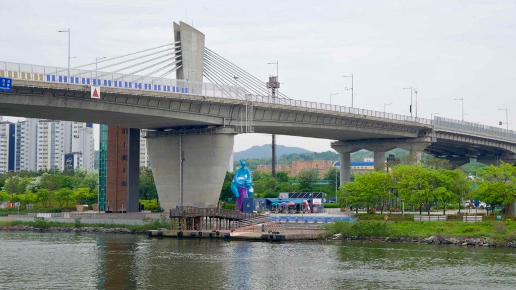 A cable-stayed span of Sicheon Bridge stretches over the Ara Waterway, while below it Sicheon Waterside Park features a colorful abstract sculpture, dock, and shaded café area surrounded by spring greenery.