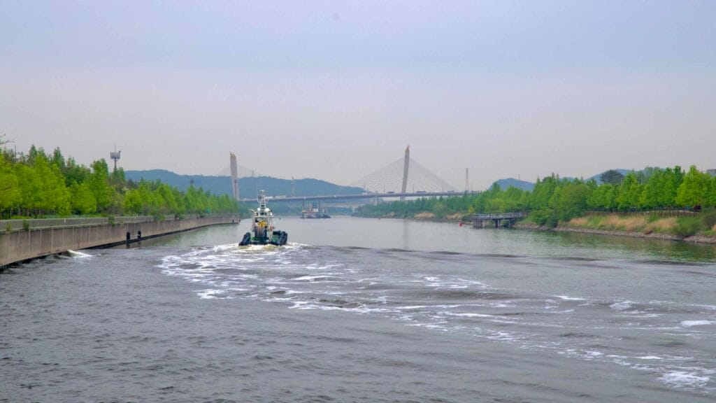 A tugboat sails east along the Ara Waterway toward the Incheon Bridge, flanked by lush greenery and canal-side infrastructure under an overcast sky.