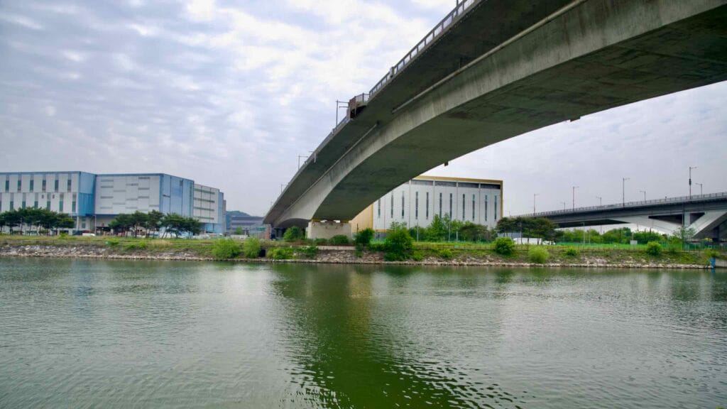 The concrete span of Hana Bridge curves over the Ara Waterway near Ara Hangang Lock, with industrial warehouses and calm canal waters below.