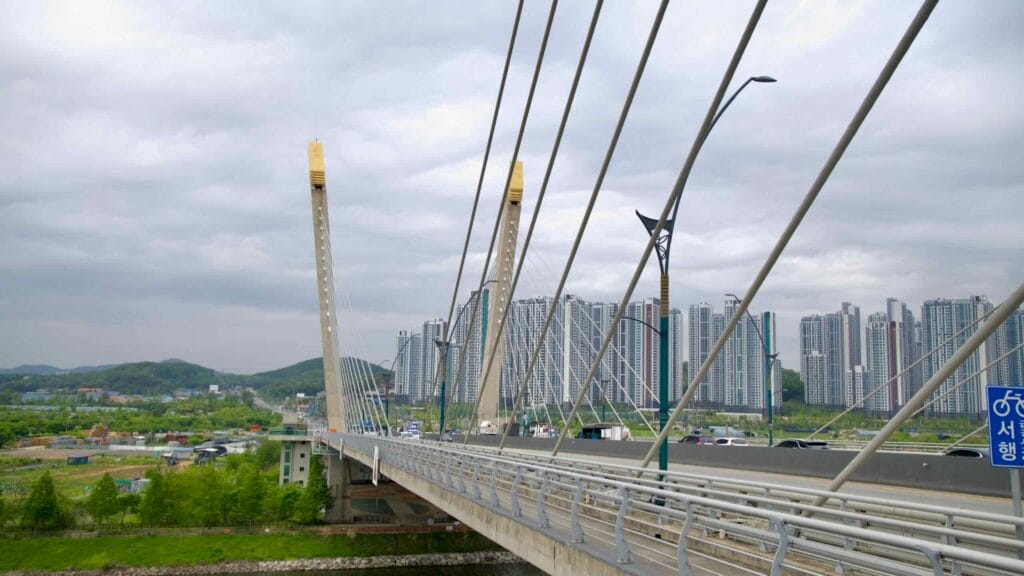 Standing atop Baekseok Bridge, a cable-stayed crossing over the Ara Waterway, looking toward Incheon’s dense skyline and surrounding hills under a cloudy sky.