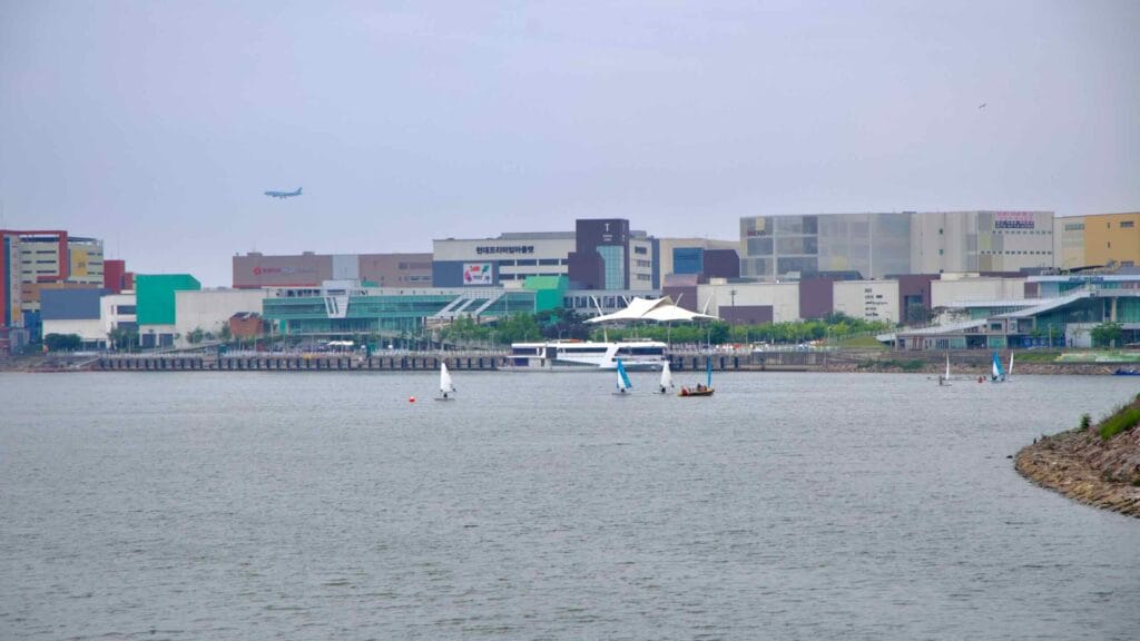 Small sailboats float near the Ara Waterway as Ara Marina and the Ara Gimpo Passenger Terminal rise in front of a dense block of logistics centers and shopping complexes near Gimpo Airport.