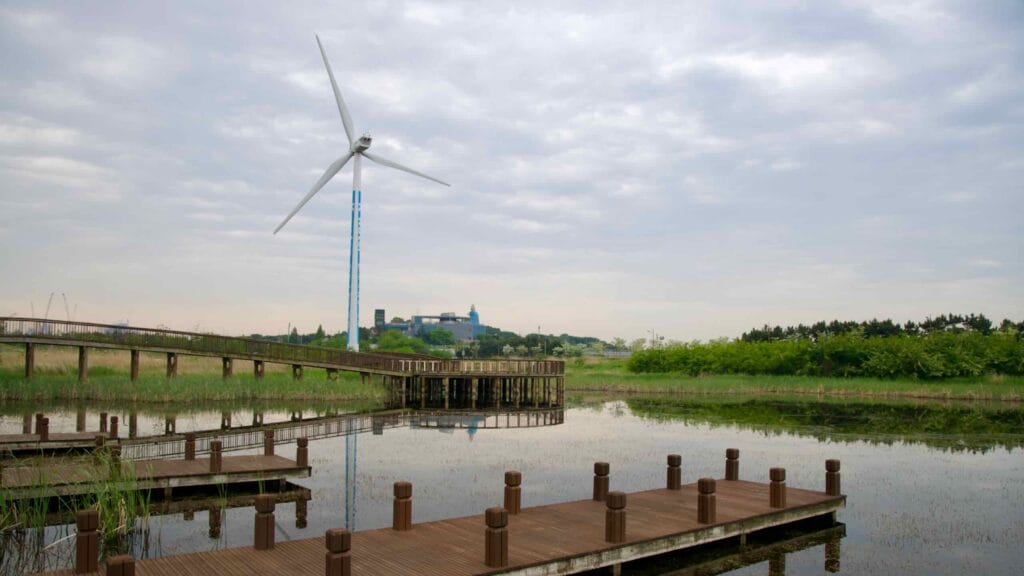 Wooden boardwalks weave through a quiet wetland at Jeongseojin, with a towering wind turbine and the Ara Waterway Environment Center visible in the background across the reflective marsh.