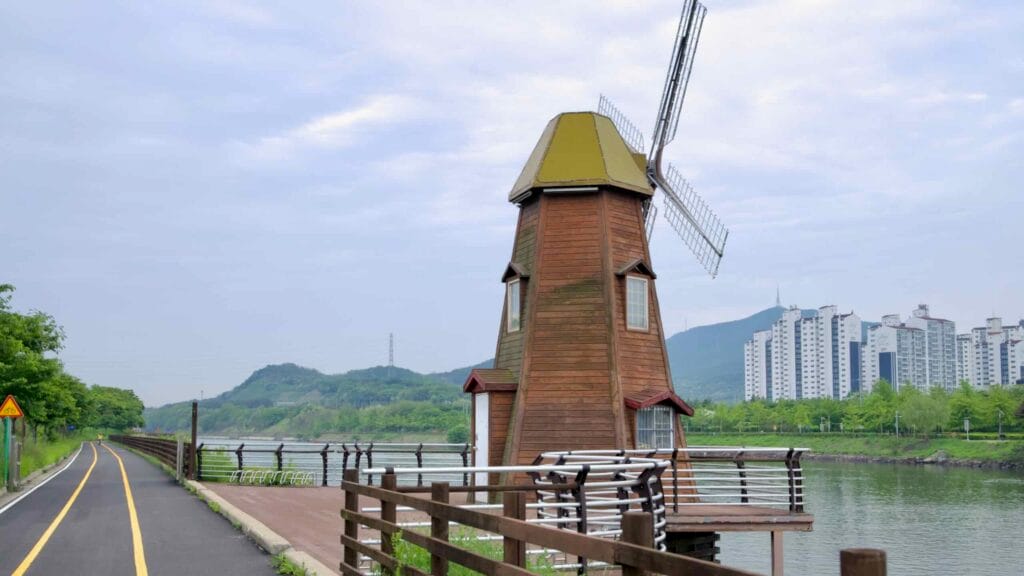 A Dutch-style wooden windmill marks a rest deck beside the Ara Canal bike path at Sicheon Waterside, with high-rise apartments across the water.