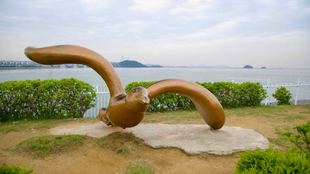 A bronze seabird sculpture with wings spread wide faces the Yellow Sea, with Yeongjong Island and Incheon Bridge visible in the misty background along the Ara Waterway coast.