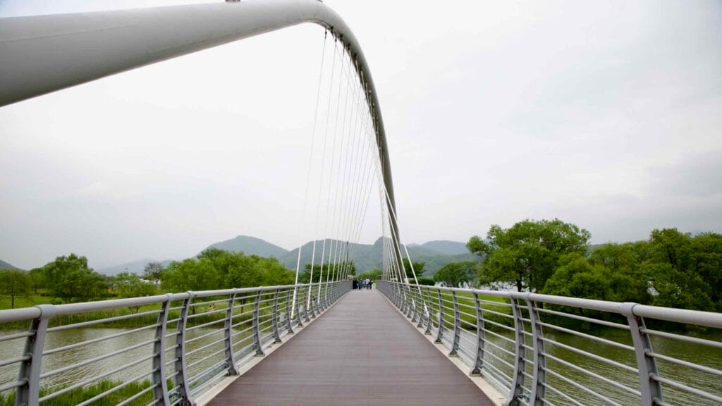 Baetnari Bridge, a curved pedestrian span, stretches over calm wetlands in Namyangju Water Garden, connecting scenic paths beneath forested mountains.