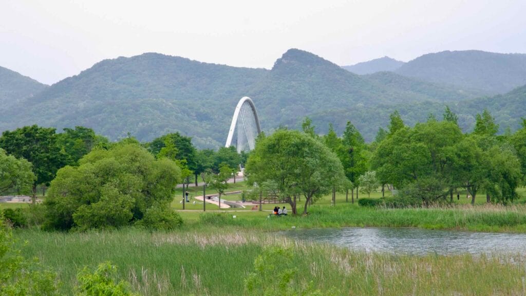 A wide view of Namyangju Water Garden shows Baetnari Bridge rising beyond the wetlands and forest paths, framed by mountains and spring greenery.