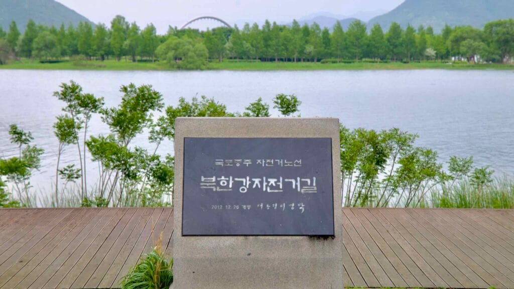 A commemorative marker for the Bukhangang Bike Path stands beside the riverbank at Balgeun Gwangjang, with Namyangju Water Garden and Baetnari Bridge in the background.