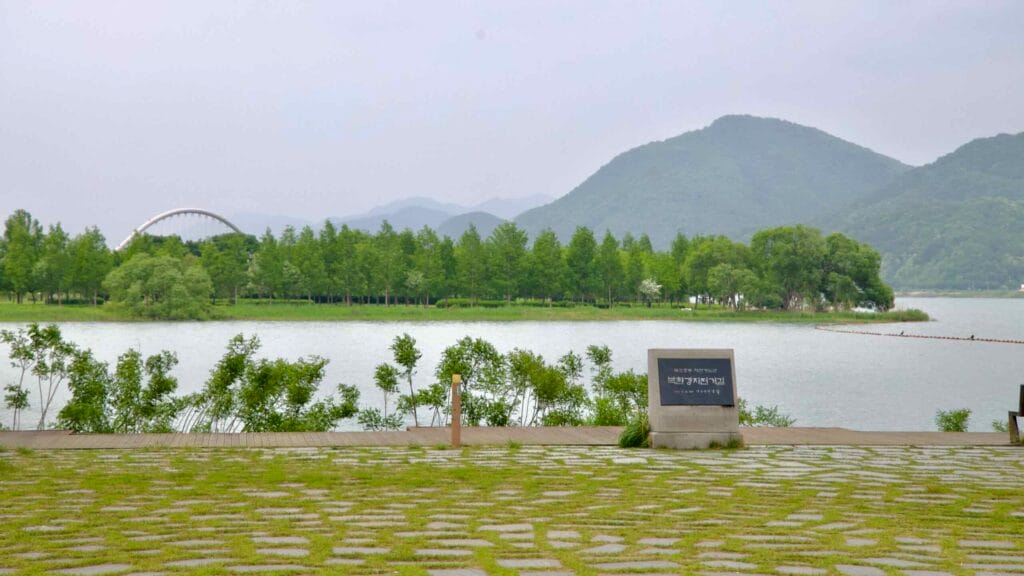 Looking across the North Han River from Balgeun Gwangjang toward Namyangju Water Garden, with Baetnari Bridge and forested hills forming a serene riverside backdrop.