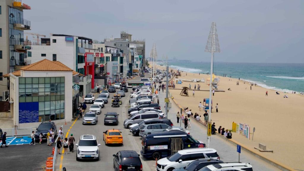 Cars fill the roadside parking and pedestrians stroll along the sandy beachfront, with shops and cafes lining the busy Gangneung Coffee Street promenade.