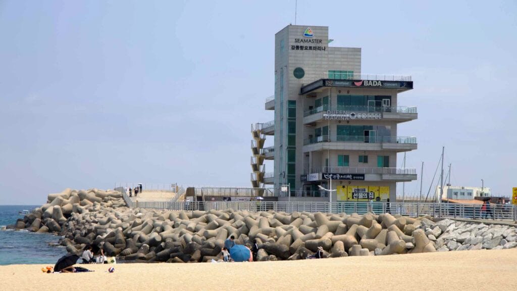 Observation tower rises behind tetrapods at Gangneung Port, with beachgoers nearby.