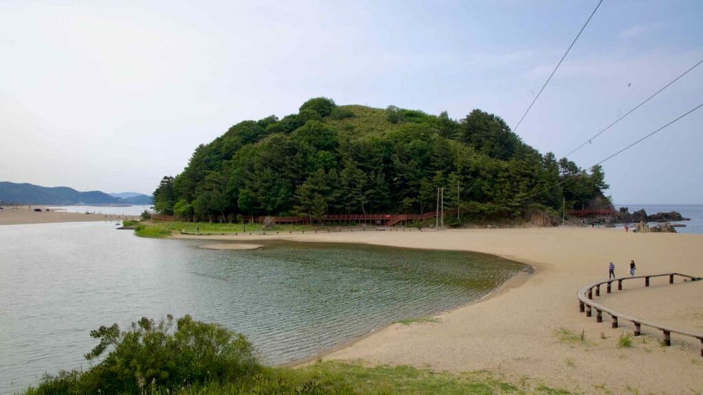 Deokbong Mountain rises beside a calm lagoon and sandy beach, with wooden walkways winding along the forested slope and shoreline near the East Sea.