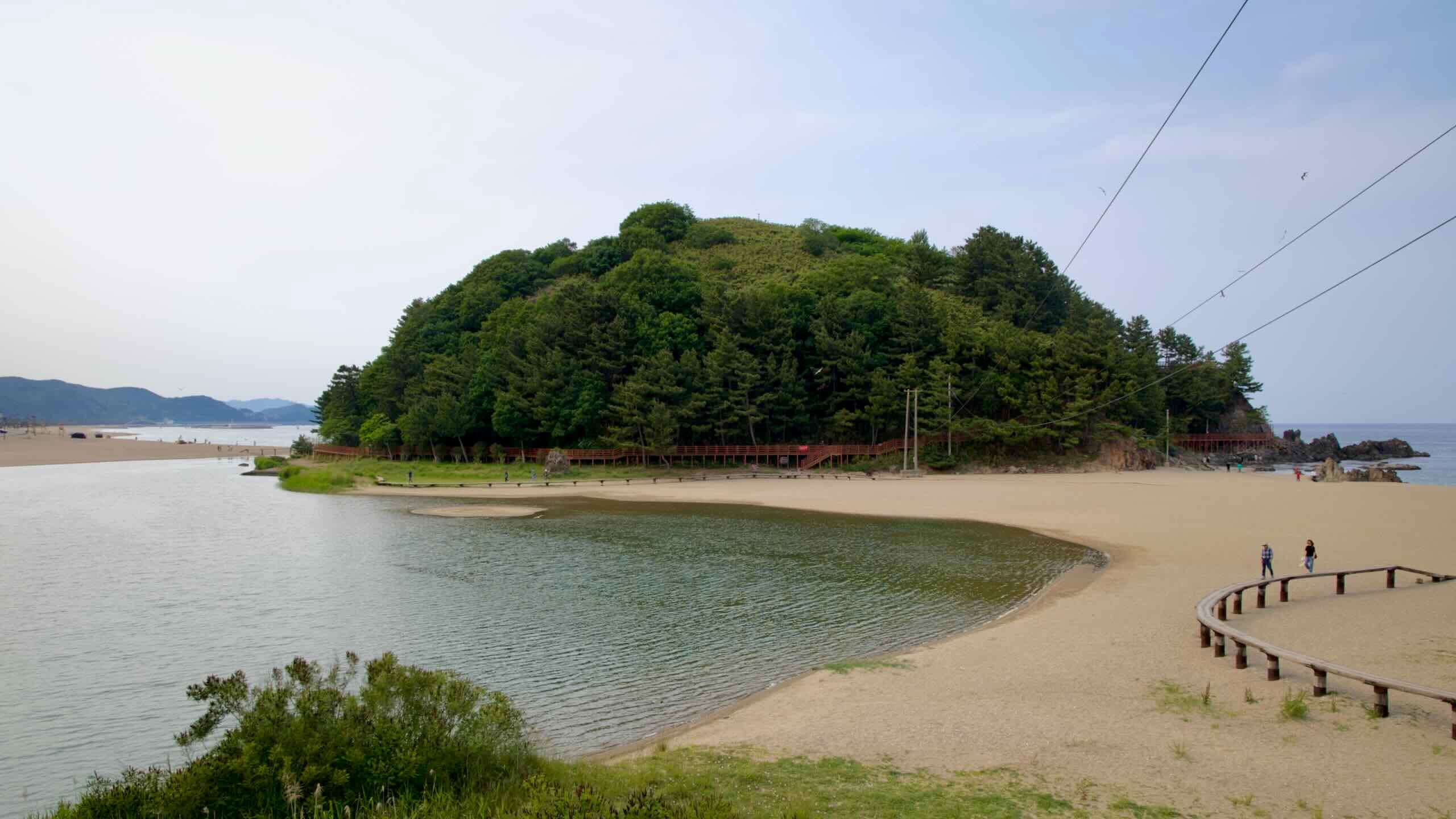 Deokbong Mountain rises beside a calm lagoon and sandy beach, with wooden walkways winding along the forested slope and shoreline near the East Sea.