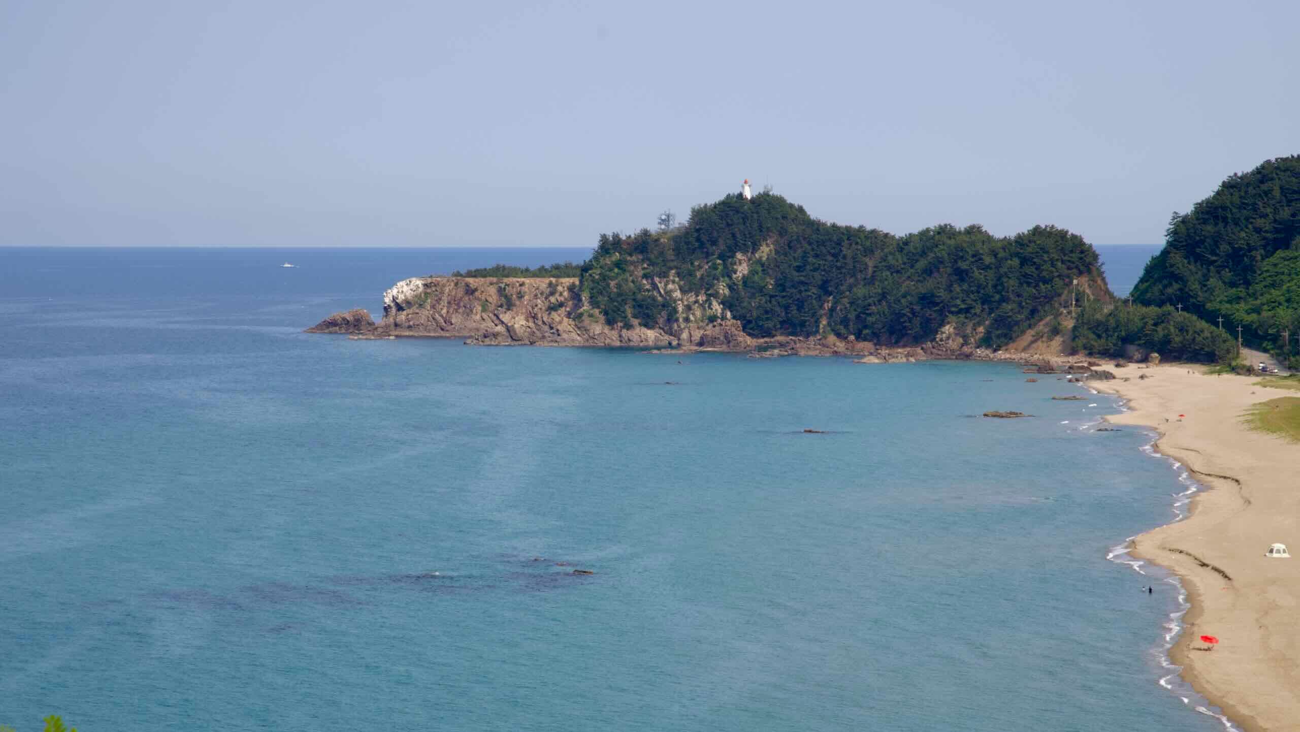 A rocky, forested cape juts into the East Sea at the edge of Deoksan Beach, with a small white lighthouse perched above and gentle waves brushing the sandy shore.