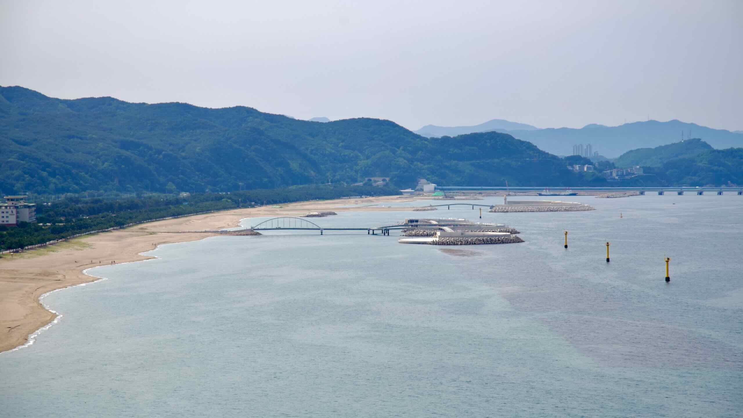 From a mountain lookout, Maengbang Beach unfolds along the coast with protective breakwaters, pedestrian bridges, and forested ridges rising in the background.