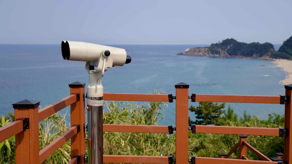 A binocular viewer atop Deokbong Mountain overlooks the East Sea and a forested headland near Deoksan Beach, offering clear views across the coastal waters.