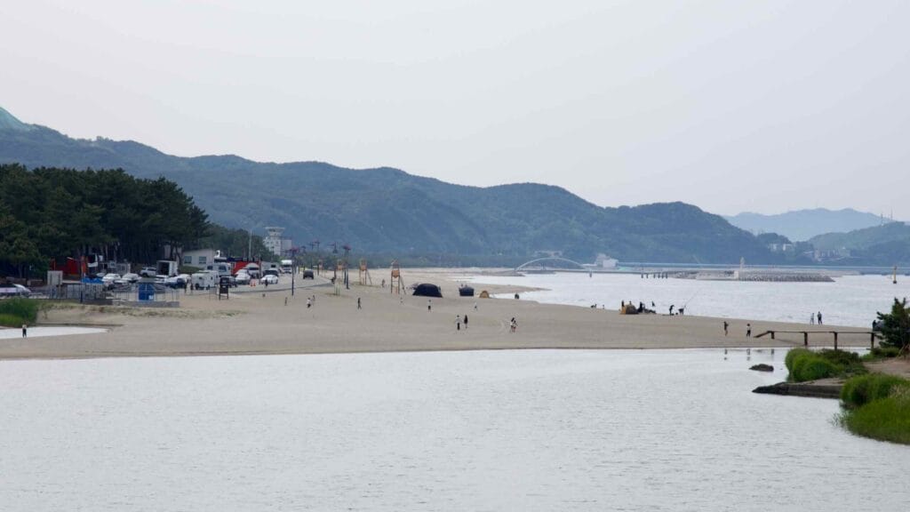 Campers and beachgoers dot the wide stretch of Maengbang Beach, as pine groves, lifeguard towers, and breakwaters line the coast under hazy mountain silhouettes.