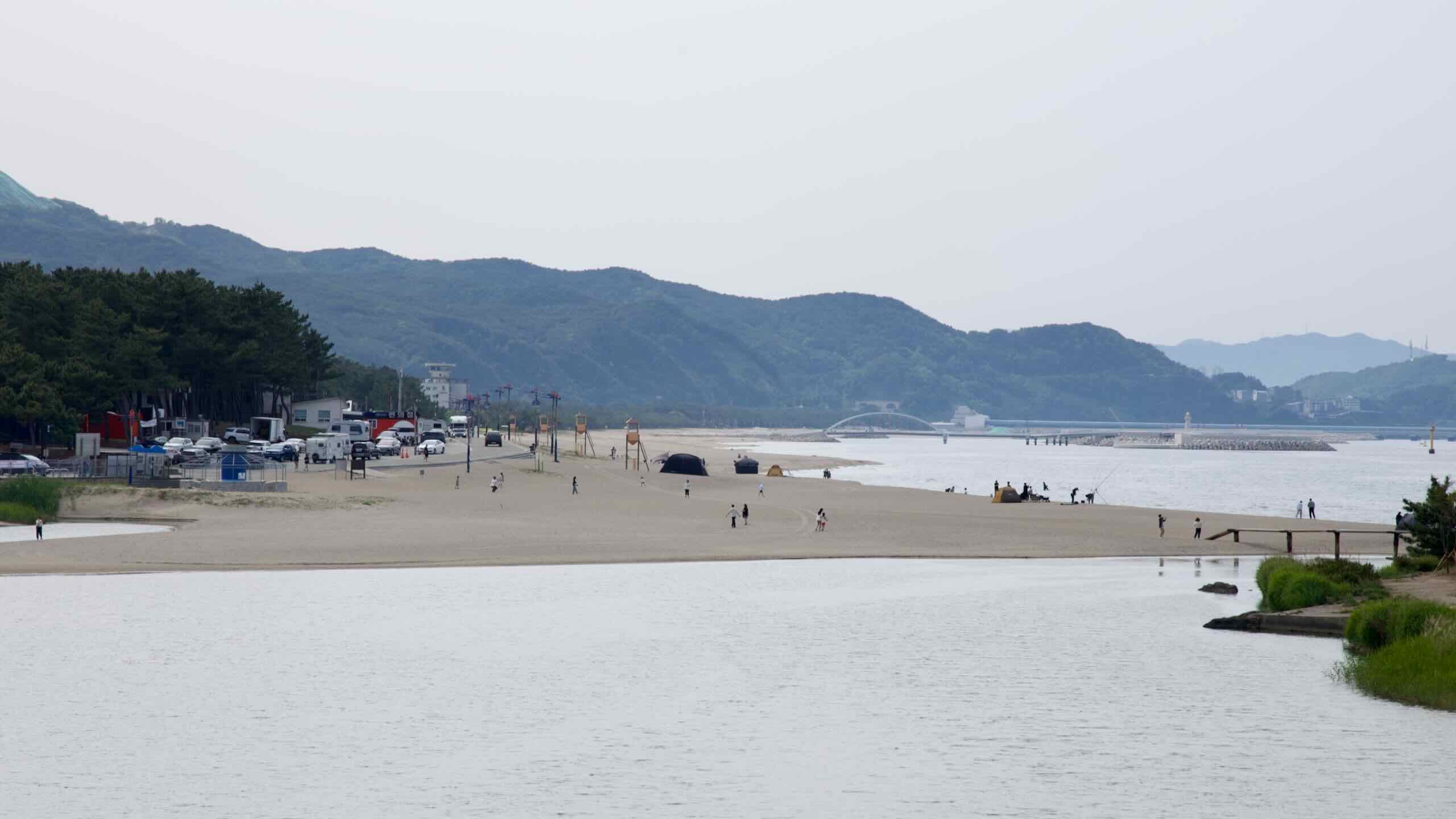 Campers and beachgoers dot the wide stretch of Maengbang Beach, as pine groves, lifeguard towers, and breakwaters line the coast under hazy mountain silhouettes.