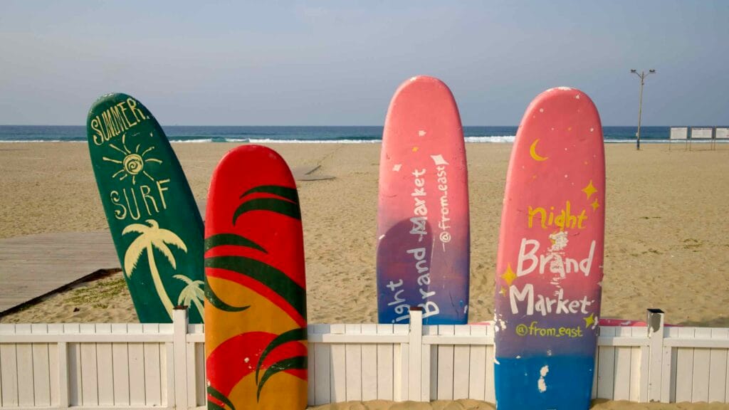 A set of painted surfboards with tropical and market-themed designs stands upright behind a white fence along the sandy shoreline of Mangsang Beach.