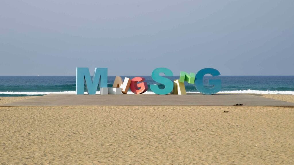 A playful sculpture spelling “Mangsang” stretches across a beach platform, set against low waves rolling onto the golden sand of Mangsang Beach.