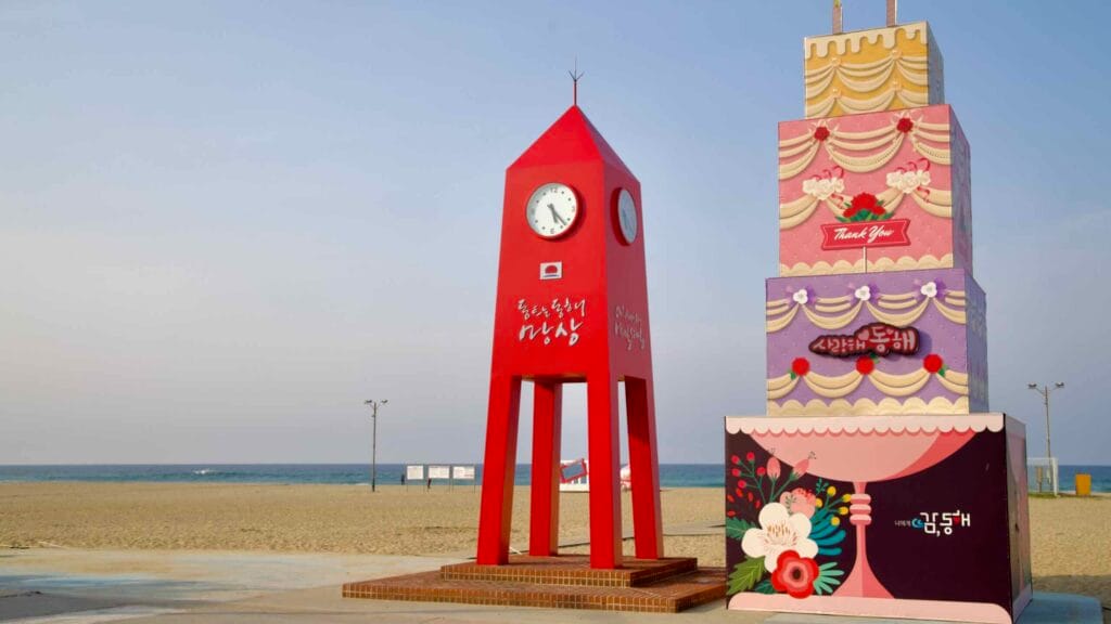 A vibrant red clock tower and tiered birthday cake monument stand near the water’s edge on Mangsang Beach, celebrating the city of Donghae in bold pastel colors.