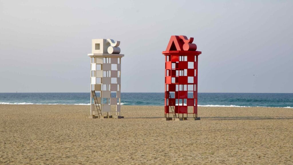Two tower-like sculptures in red and white checkerboard patterns stand side by side on the sands of Mangsang Beach, facing calm waves of the East Sea.