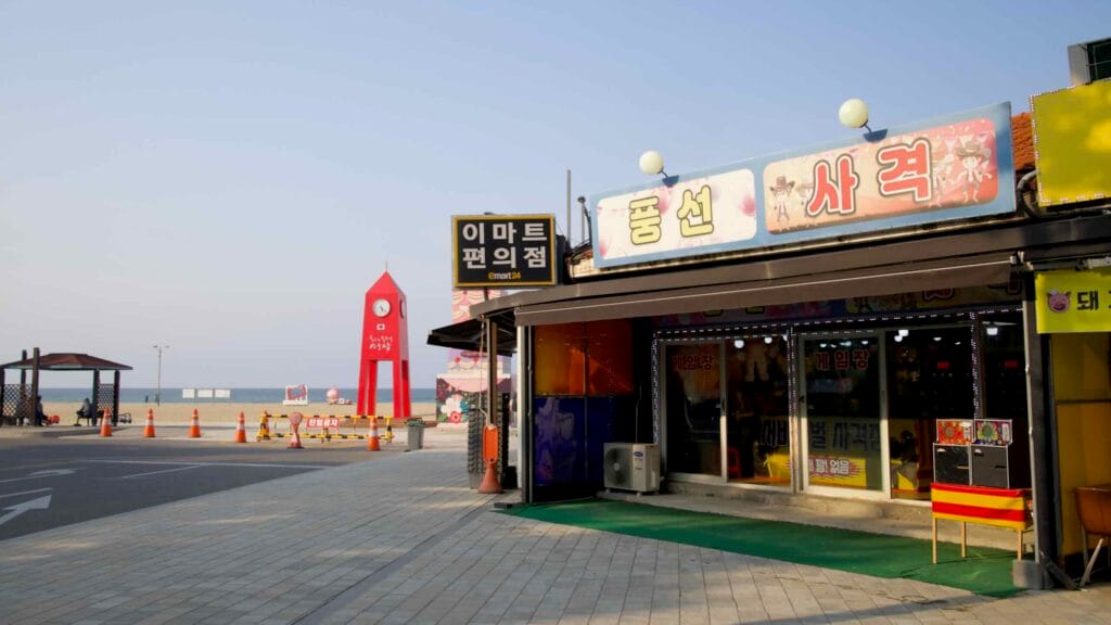 A convenience store and balloon shooting game stall sit beside the iconic red clock tower at Mangsang Beach, where roads meet sand and the East Sea begins.