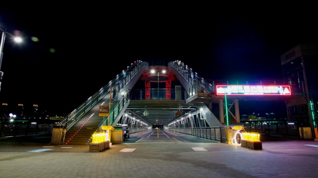 The illuminated ramp and elevator of Geumgang Pedestrian Bridge rise above an empty riverside plaza.