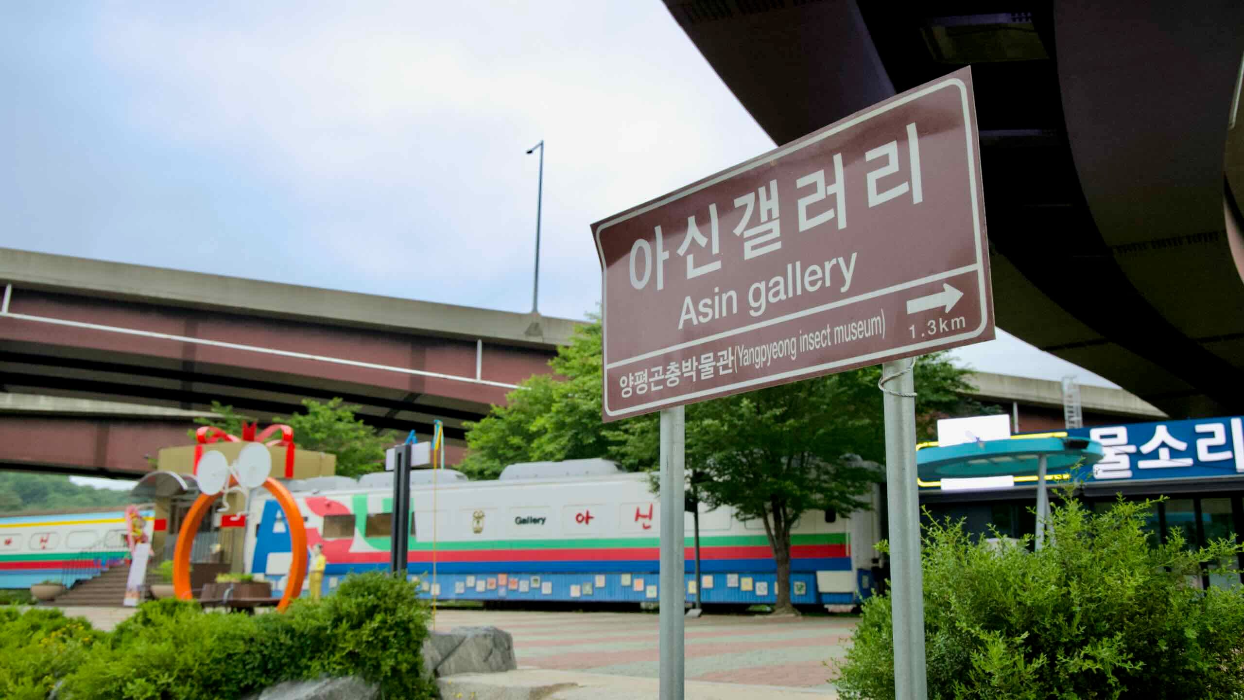 A brown road sign points 1.3 km to Asin Gallery at the repurposed Asin rail station, beneath an overpass beside a colorful train-car art museum.