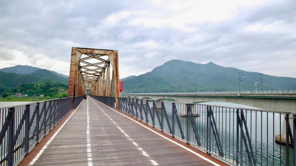 A cyclist enters the rusting frame of the old Bukhangang Railroad Bridge, now a riverside bike path with forested peaks rising in the background.