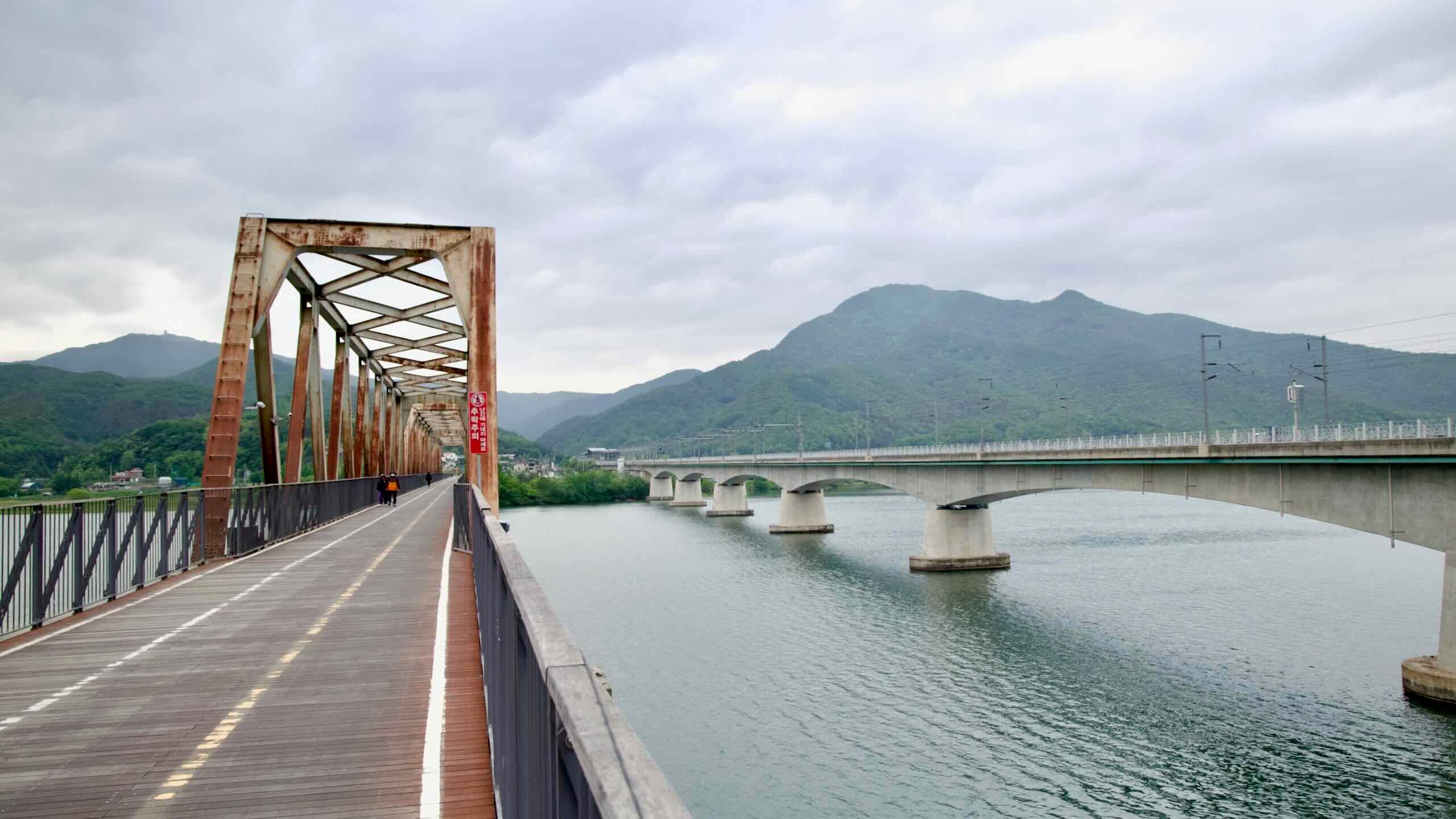 The old rusted Bukhangang Railway Bridge parallels the modern Yangsu Railway Bridge, both spanning the North Han River beneath forested mountains.