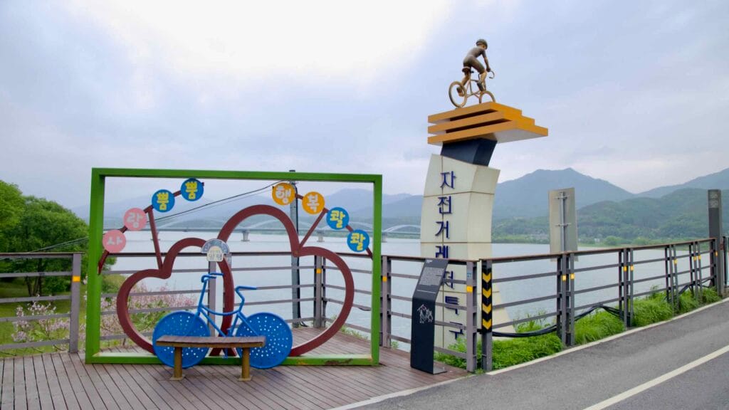 A cyclist statue and colorful heart-shaped photo zone greet riders at the entrance to the Bukhangang Bridge bike path overlooking the North Han River.