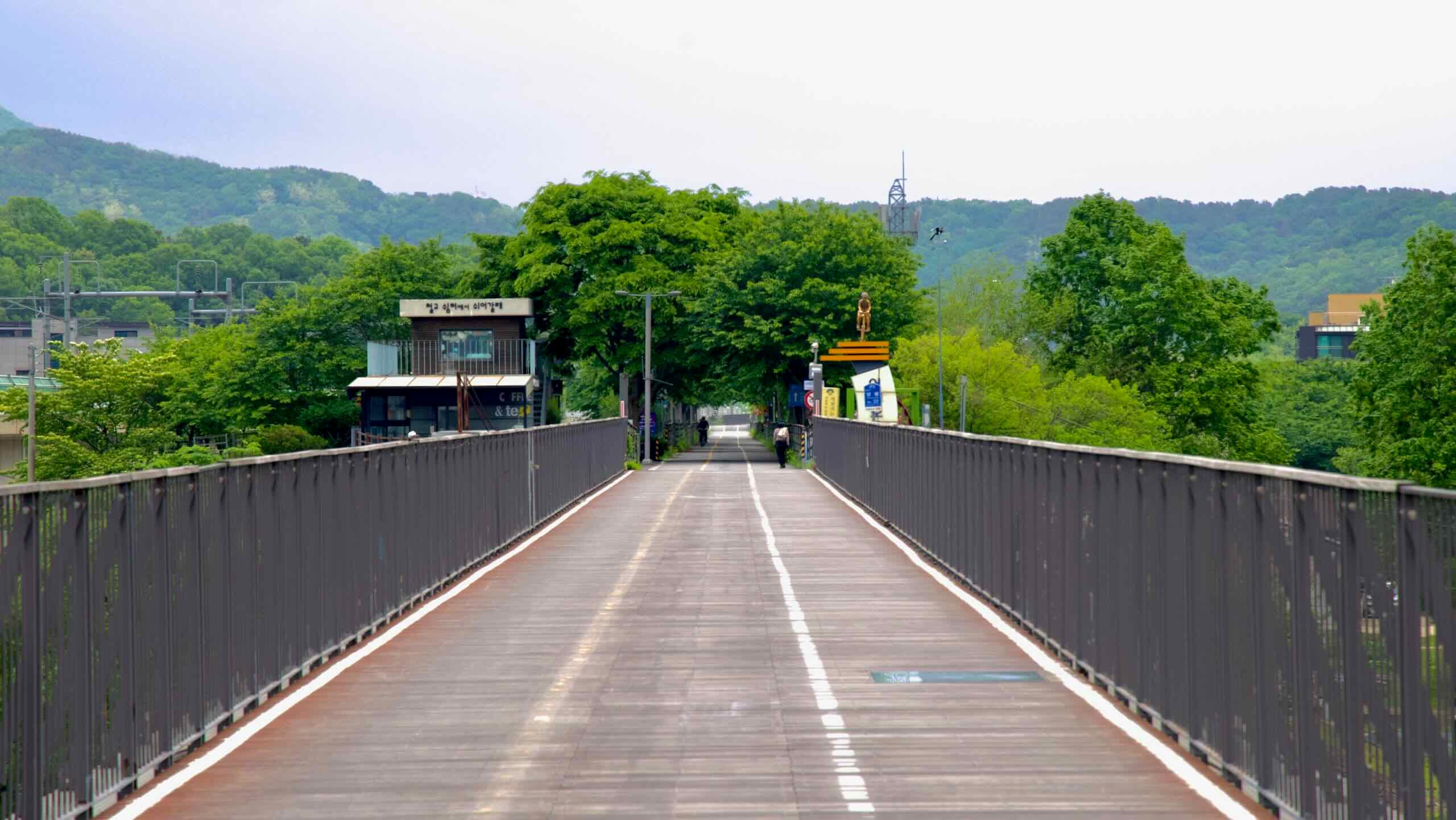 The Bukhangang Railway Bridge ends in a green corridor of trees, cafés, and bike facilities, with a gold cyclist statue marking the start of the riverside trail.