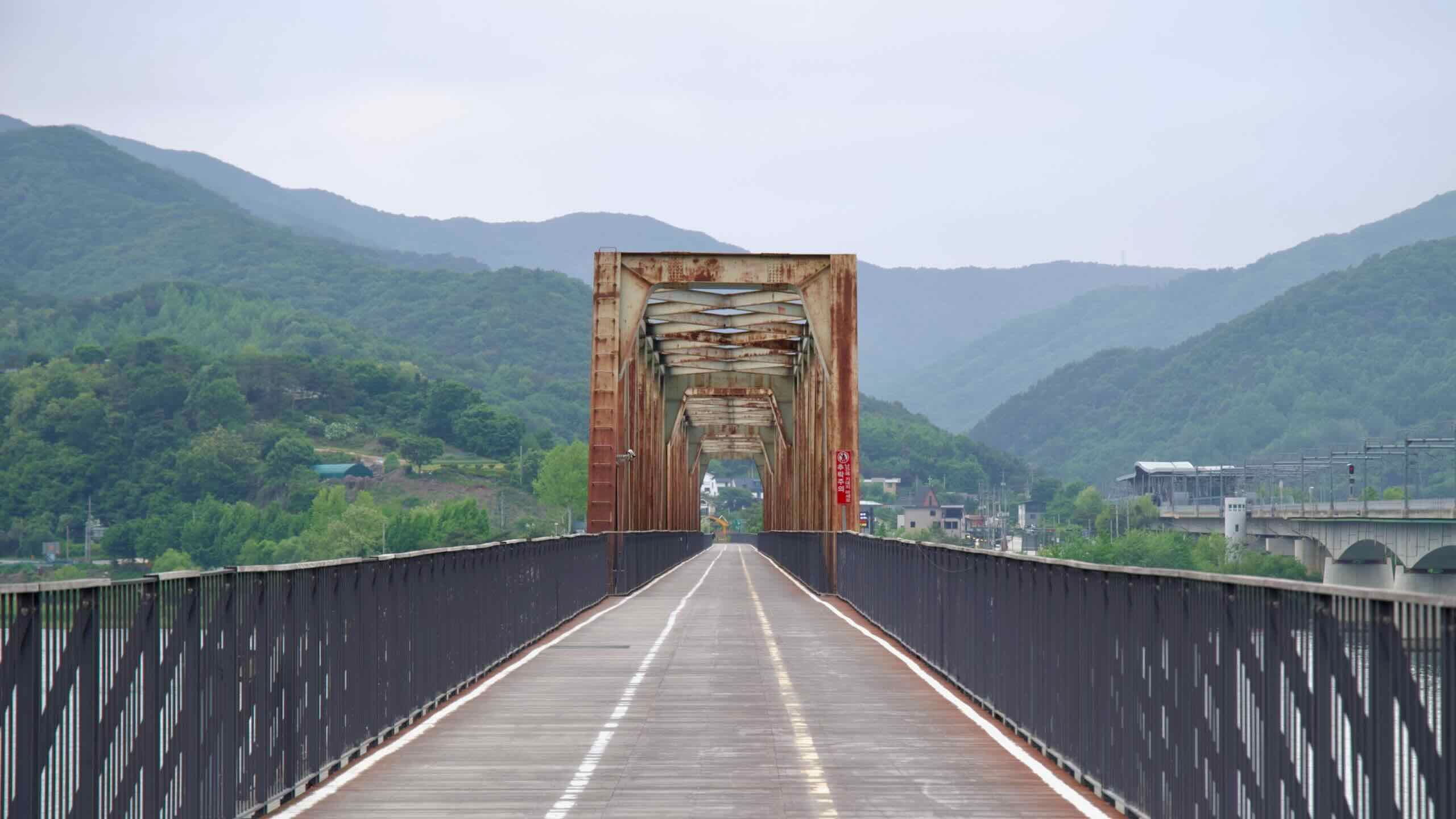The rusted trusses of the Bukhangang Railway Bridge stretch toward the green hills and distant Yangsu Station, now serving cyclists on Korea’s scenic Hangang path.