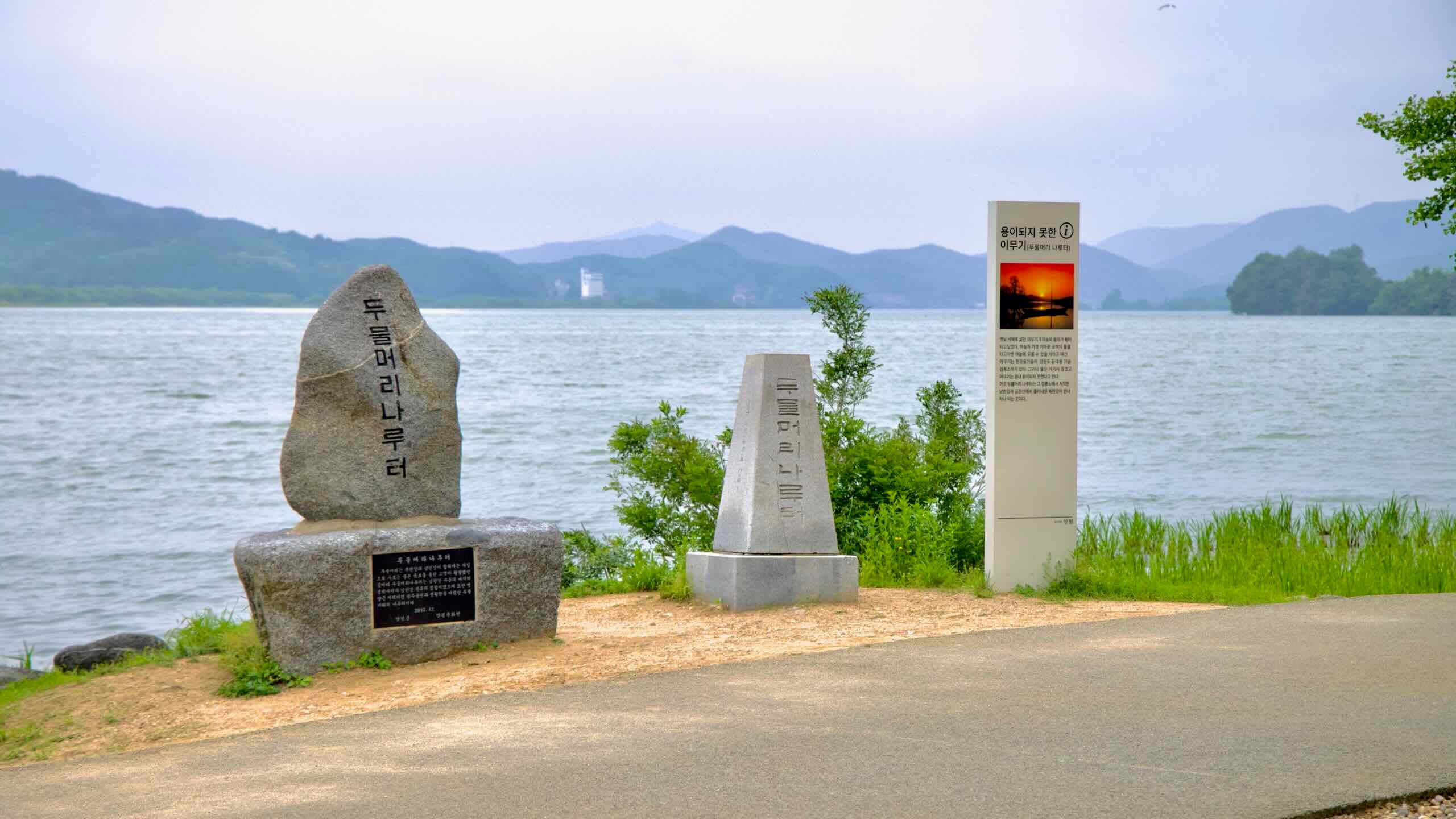 Stone markers and an informational sign stand along the riverbank at Dumulmeori, a scenic spot where the North and South Han Rivers converge.