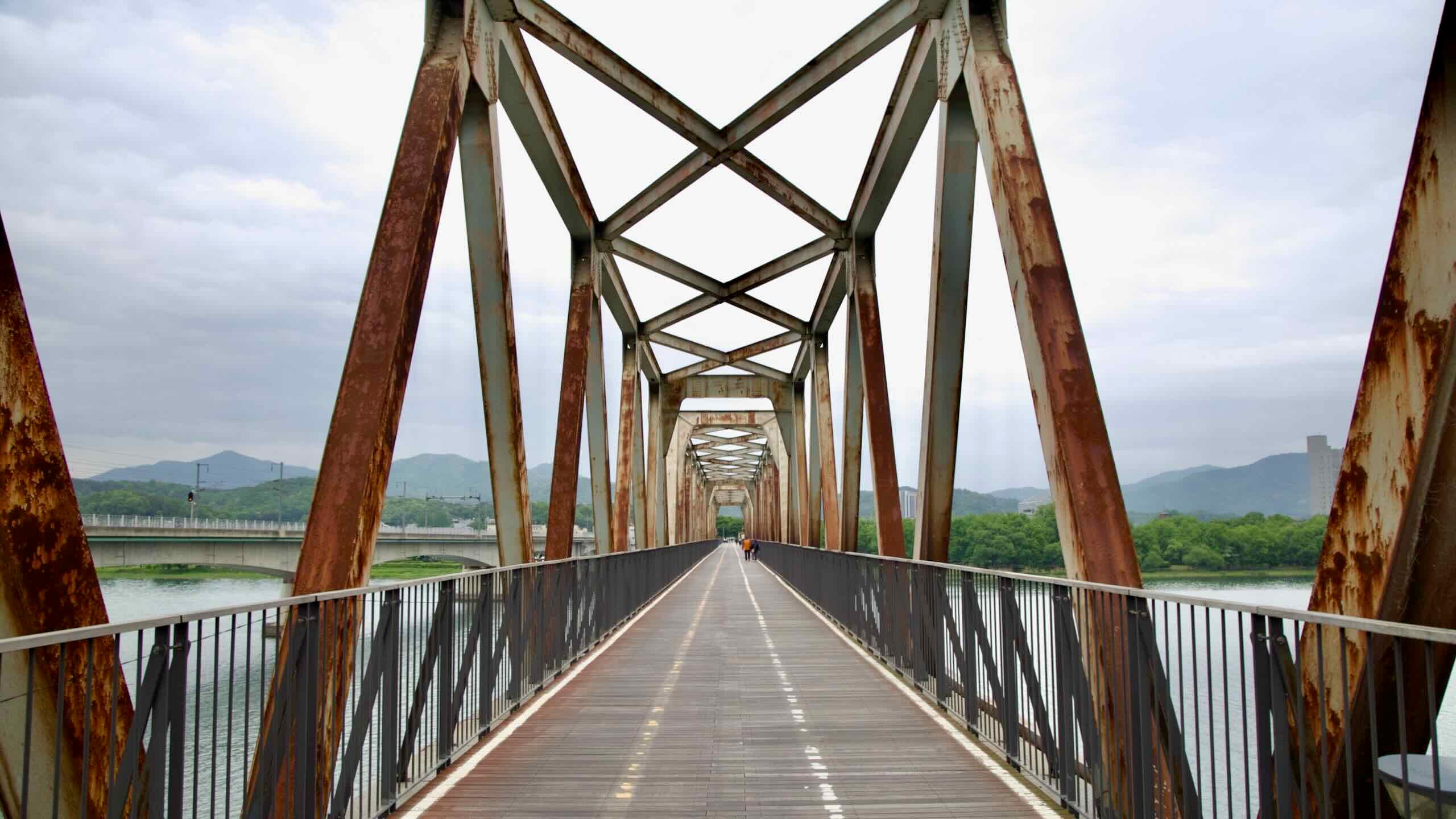 Rusted steel beams frame the old Bukhangang Railroad Bridge, now repurposed as a bike and pedestrian path above the North Han River.