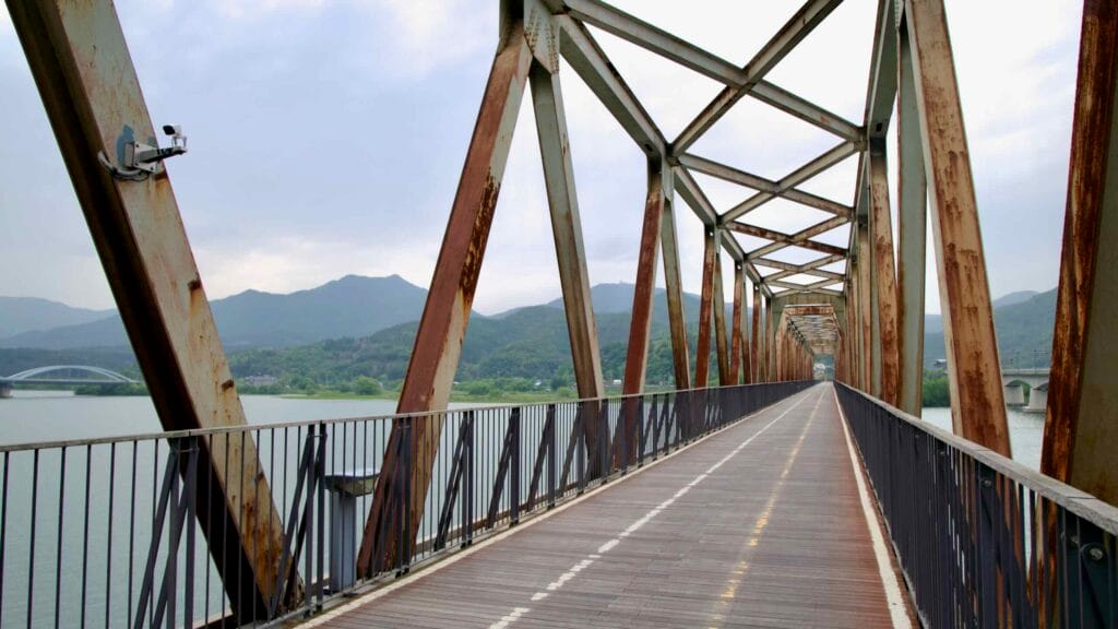 The Bukhangang Railway Bridge reveals rusted steel trusses and a straight bike path stretching across the river toward distant green hills.
