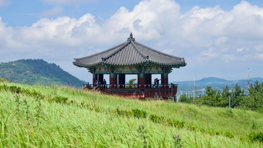 Suwol Pavilion stands above windy meadows with oreum ridges beyond.