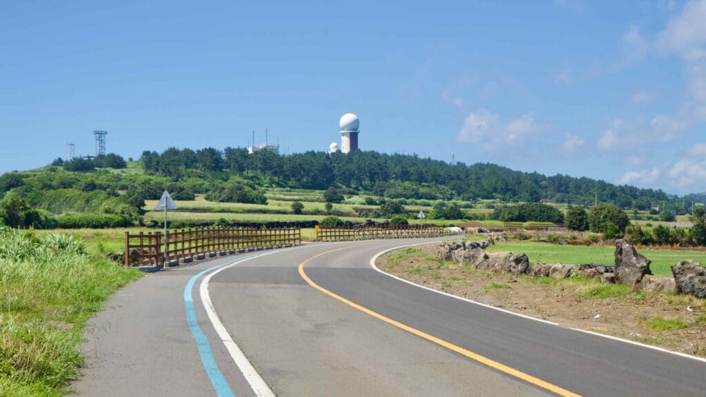 Blue‑striped road bends toward hills with radar domes near Suwolbong, Jeju.