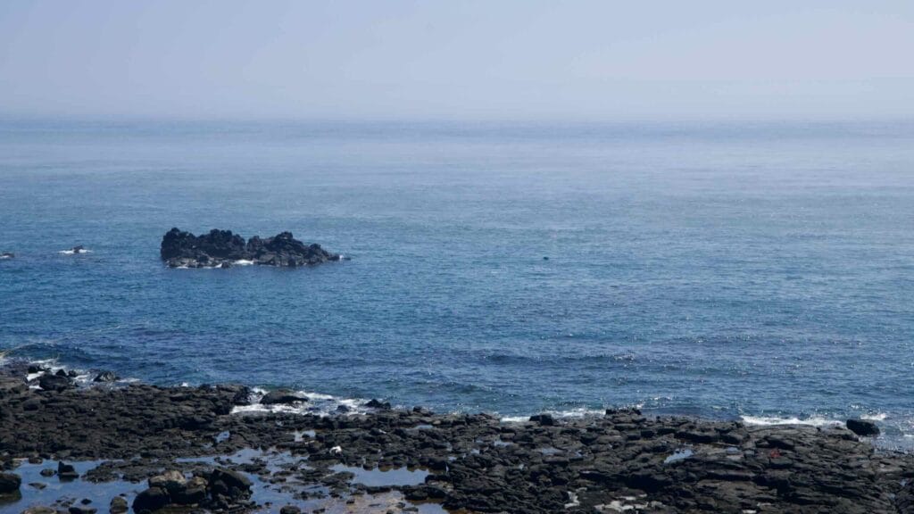 Basalt islet off Jeju’s west coast beyond a lava‑rock shoreline.