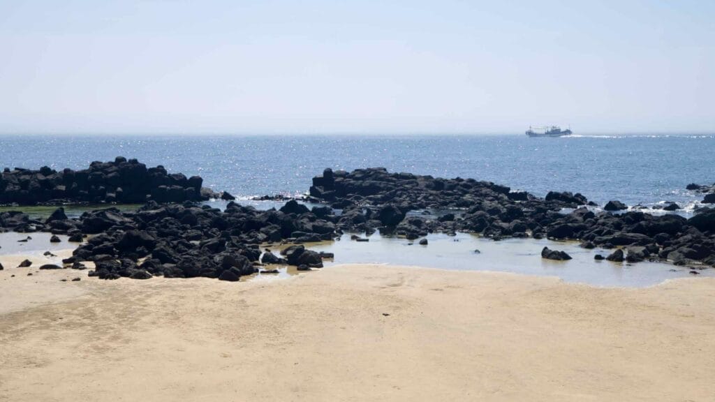 Sandy cove with lava rocks and a distant fishing boat on Jeju’s west coast.