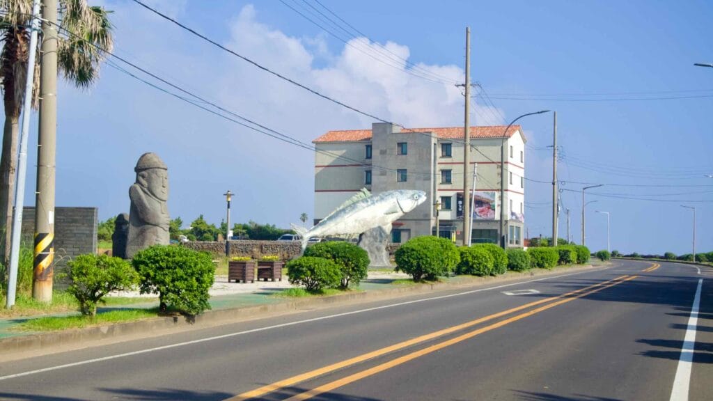 Dol hareubang and large fish sculpture beside a coastal road.