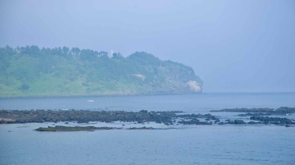 Songaksan’s forested headland rises above calm water with dark basalt reefs.