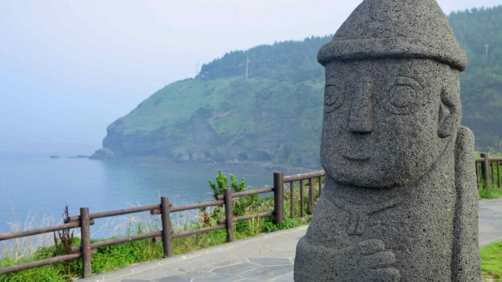 A dol hareubang stone guardian stands beside the cliff‑top rail above the calm bay at Songaksan.