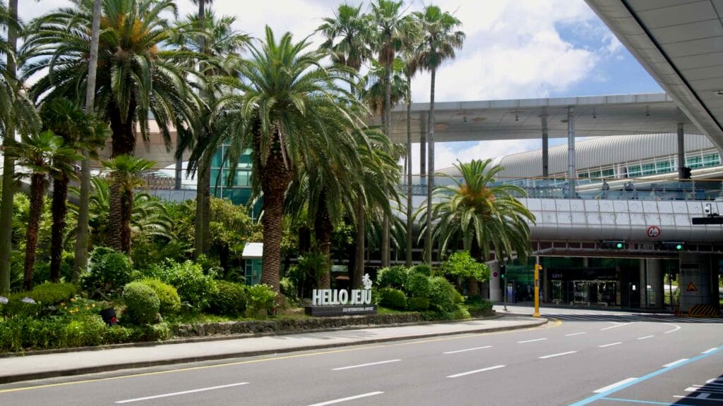 Palm‑lined entrance and “HELLO JEJU” sign outside Jeju International Airport.