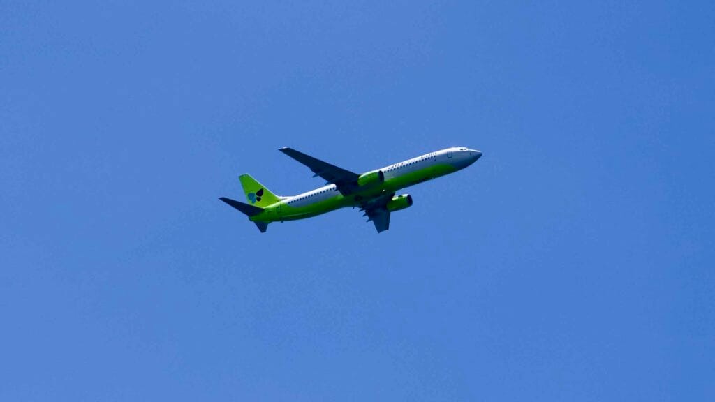 Green‑tailed airliner climbs above Jeju City under a blue sky.