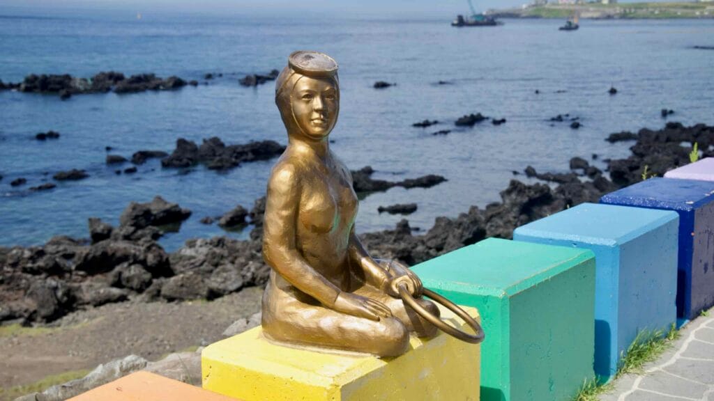 Bronze haenyeo statue on a rainbow seawall, gazing out over Jeju’s black lava coast.