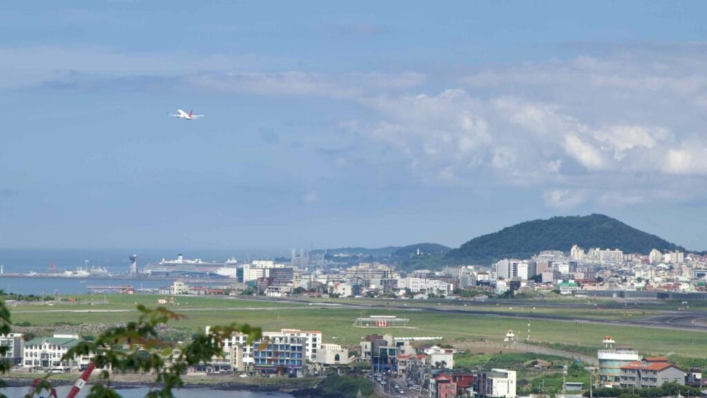 Departure shot—aircraft banking over the coast with low hills and harbor beyond.